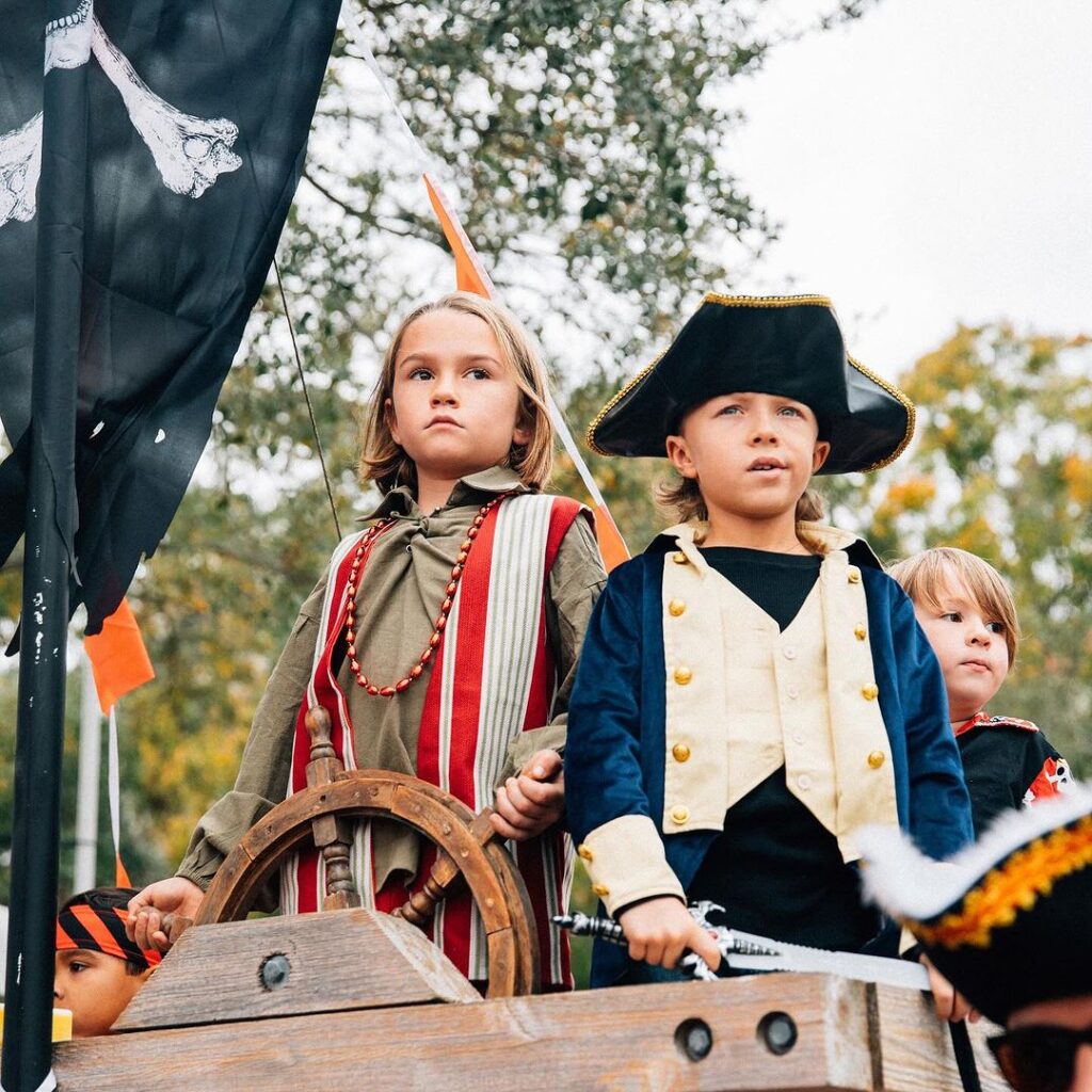 A group of children dressed in elaborate pirate costumes stand by the waterfront in Downtown Tampa, with one little girl in focus, gazing wistfully towards the horizon as palm trees and the city skyline form a backdrop, capturing the youthful excitement of the Children’s Gasparilla Festival.