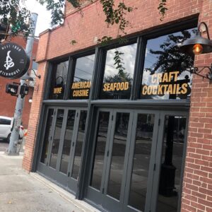 The exterior of Barterhouse Ybor, with its classic brick facade, presents a welcoming entrance on a lively street in Ybor City. The window decals boast the offerings inside: "Wine," "American Cuisine," "Seafood," and "Craft Cocktails," signaling the variety and quality of the gastronomic experience that awaits within. The restaurant's name and the historical significance of the neighborhood, established in 1885, are proudly displayed, inviting patrons to step into a space where Tampa's past and present culinary delights converge.