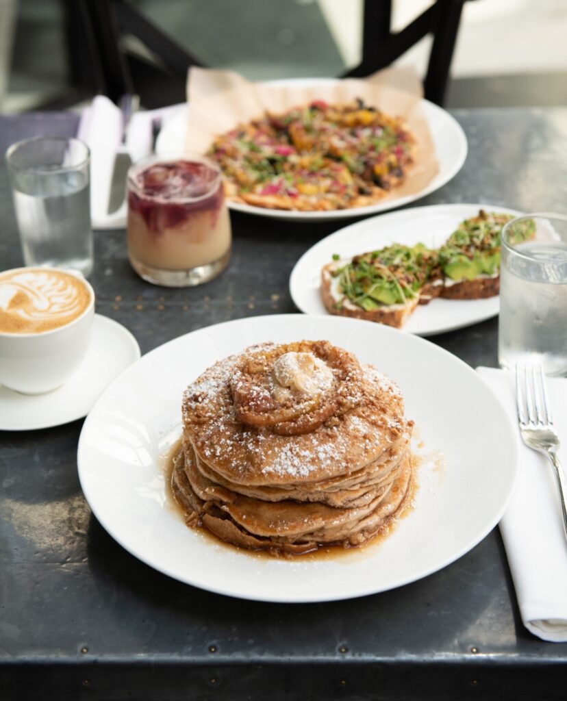 A delicious brunch spread at a Downtown Tampa cafe, featuring a stack of fluffy pancakes topped with a pat of butter and powdered sugar, accompanied by latte art coffee, avocado toast, and a fresh fruit yogurt parfait, embodying the best brunch in Downtown Tampa