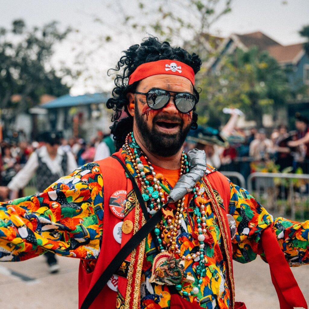 A lively participant at the Gasparilla Parade in Downtown Tampa, dressed in a flamboyant pirate costume with a red headband, smiles enthusiastically amidst the festivities.