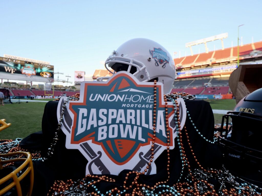 A football helmet adorned with colorful beads sits in a stadium, featuring a sign for the "Union Home Mortgage Gasparilla Bowl," with the event logo prominently displayed, all set against the backdrop of Raymond James Stadium, home to Tampa's Gasparilla Bowl, a highlight of the city's festive Gasparilla season.