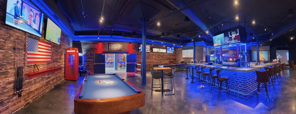 A spacious and trendy sports bar in Downtown Tampa featuring pool tables, a shuffleboard, a vibrant bar area with blue lighting, and multiple large screen TVs showcasing sports. A British and American flag complement the lively ambiance, perfect for enjoying the best brunch in Downtown Tampa.